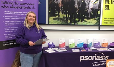A young woman with blonde hair holding a clipboard and smiling at the camera. She is wearing a purple hoodie with the Psoriasis Association logo on it, jeans and white trainers. She is standing to the left of a table full of leaflets.